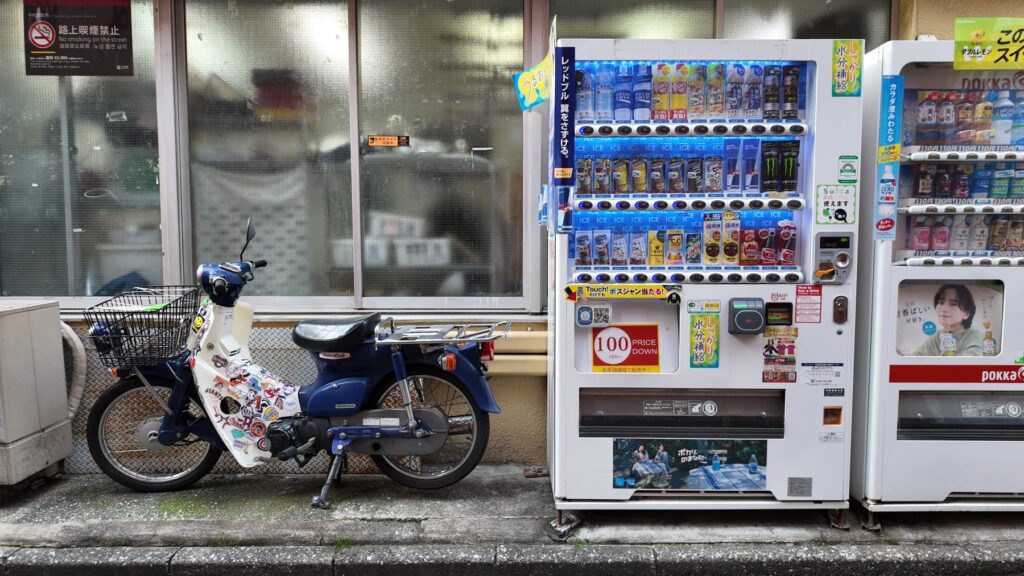 A sticker-covered Honda Super Cub parked beside glowing vending machines on an Akihabara side street